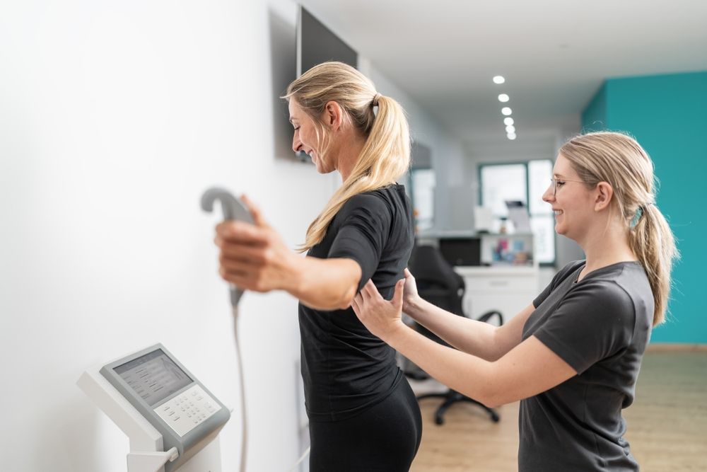 Two women exercising with a cable machine in a bright gym, one assisting the other with a stretch