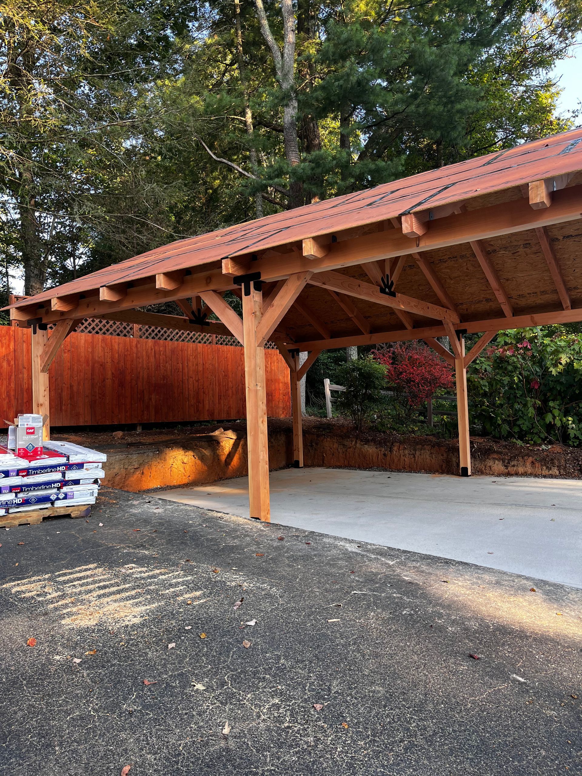 A wooden carport with a red roof is in the middle of a driveway.