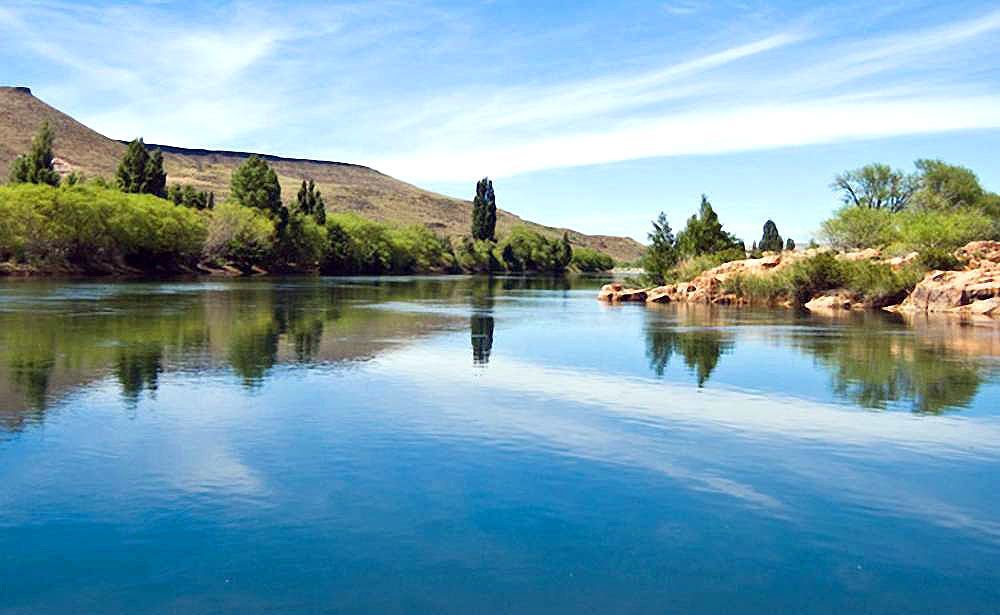 Un tranquilo lago azul refleja colinas, rocas y un cielo despejado en un día soleado.