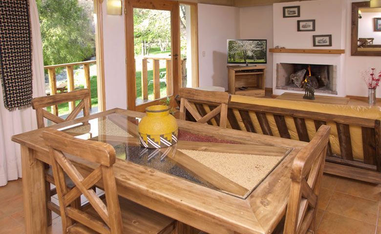 Sala de estar rústica con mesa de madera, jarrón amarillo, chimenea y televisor cerca de las puertas abiertas del patio.