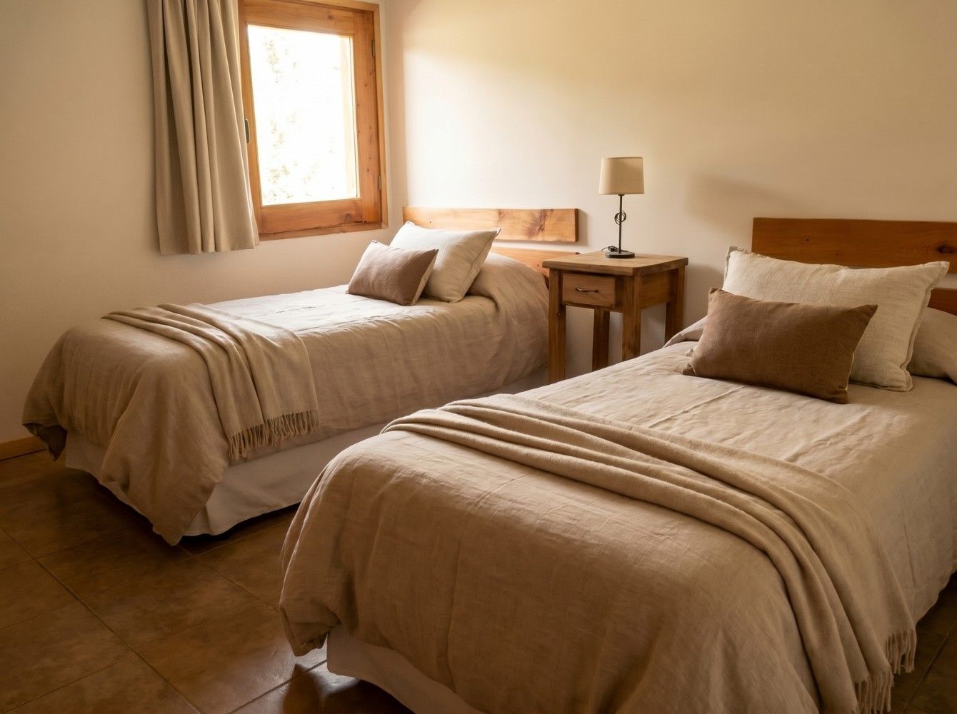Dos camas individuales bien hechas en una habitación de tonos cálidos con ventana y lámpara de noche.