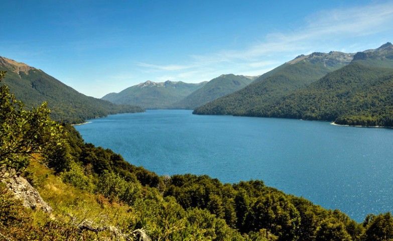 Lago de montaña azul rodeado de verdes colinas y bosques bajo un cielo despejado.