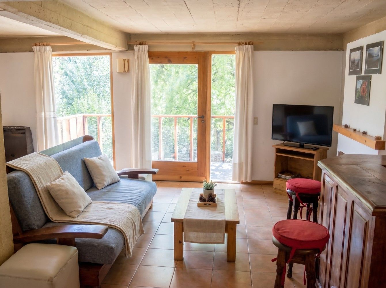 Acogedora sala de estar con sofá gris, mesa de centro, televisor, taburetes rojos y puertas corredizas de vidrio que dan a un patio.