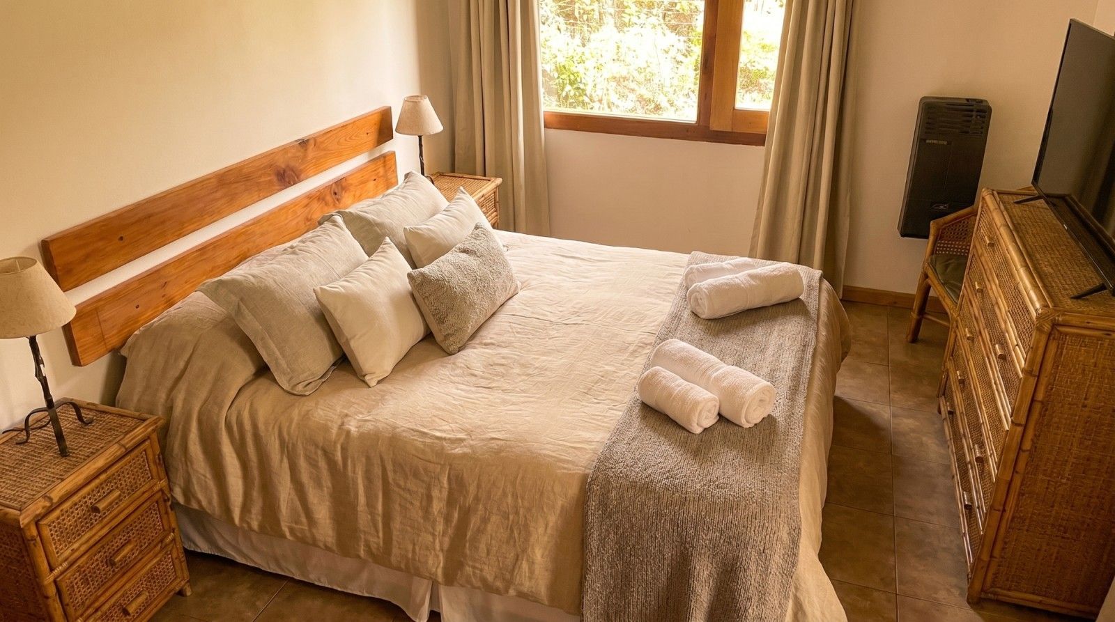 Cama bien hecha con sábanas blancas y toallas enrolladas en una habitación soleada con muebles de mimbre.