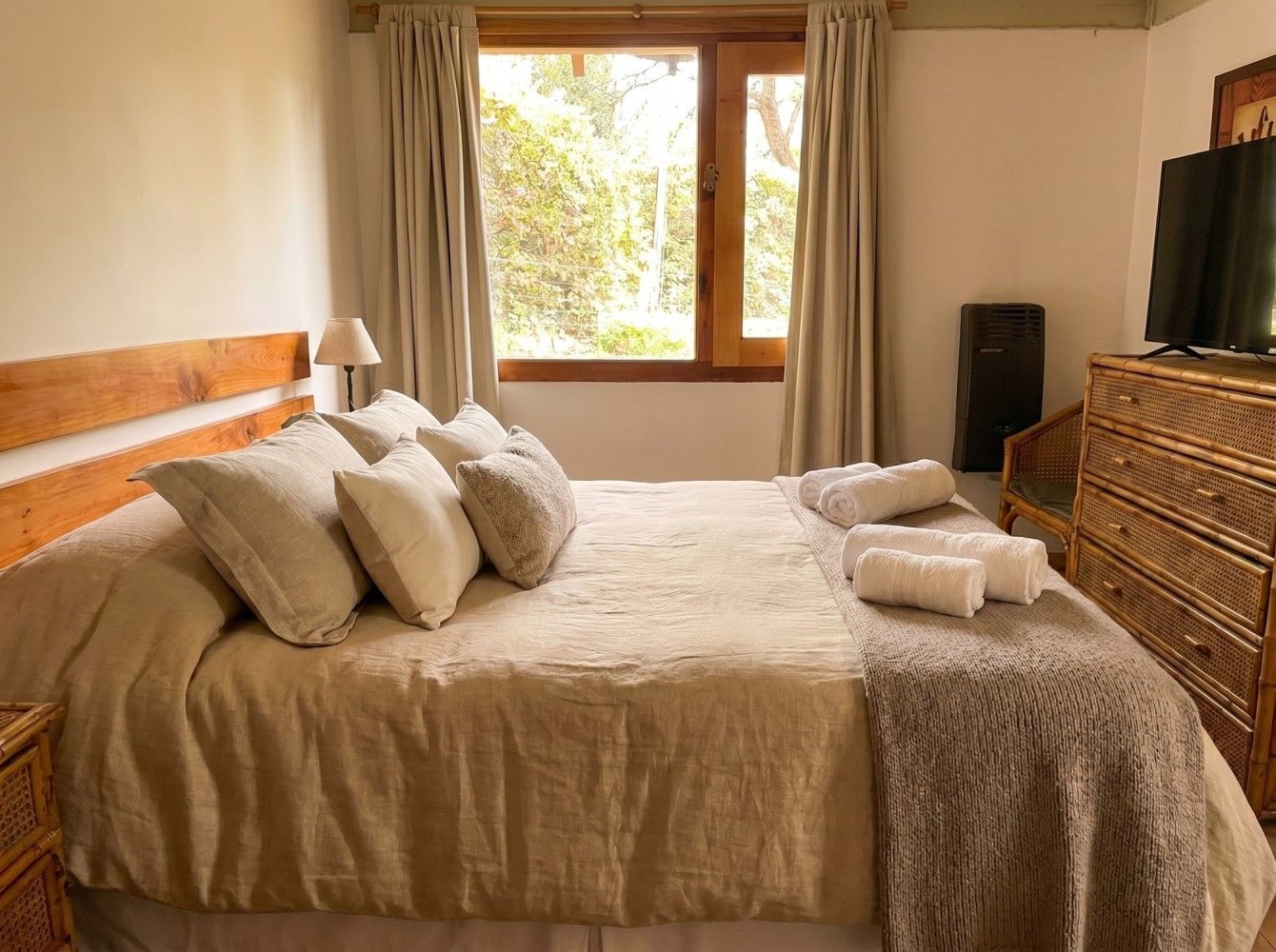 Habitación acogedora con cama beige, almohadas, ventana y cómoda de madera con televisor.