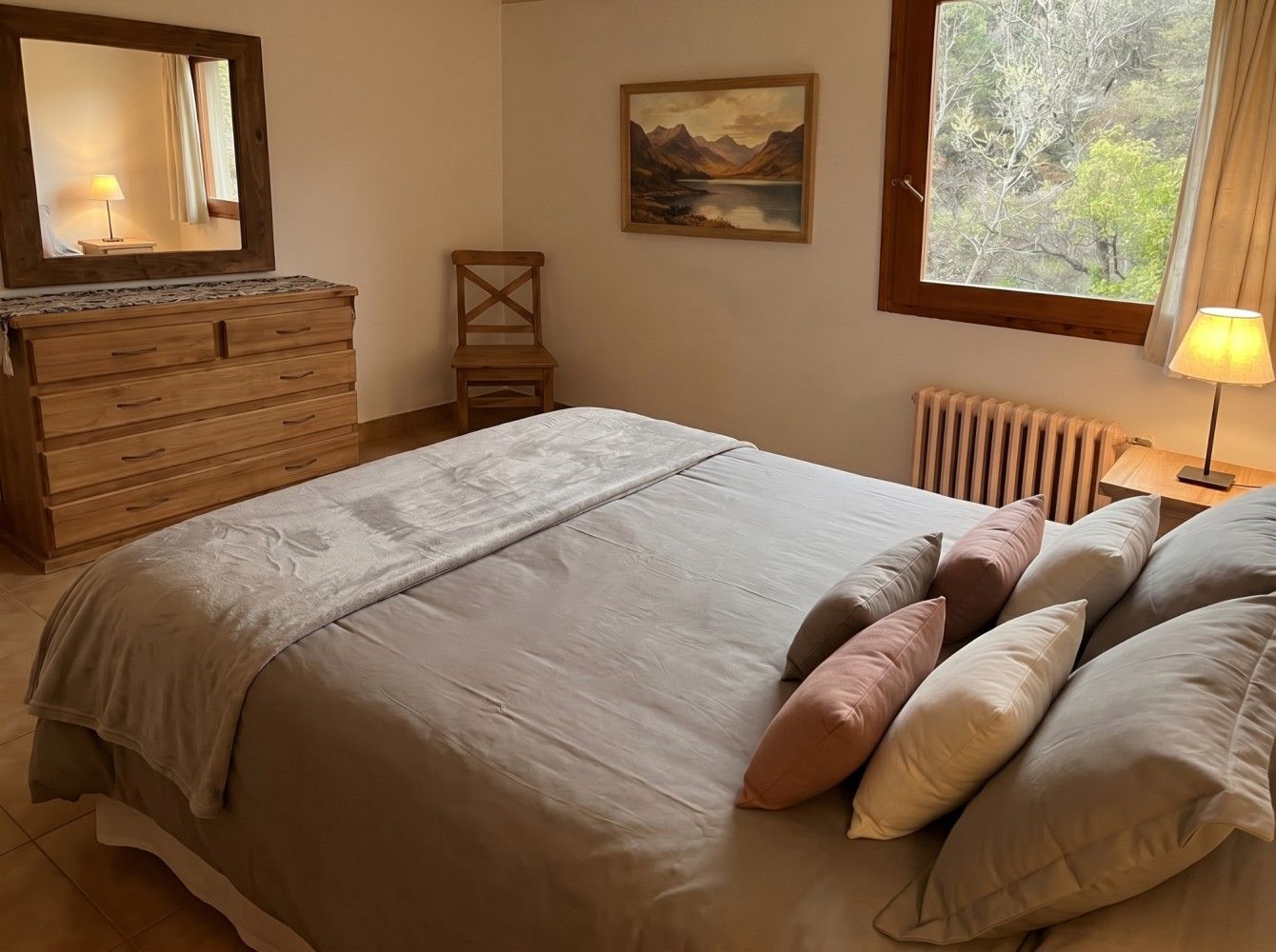 Acogedora habitación con cama hecha, cómoda, silla, lámpara, espejo y ventana con vistas a árboles.