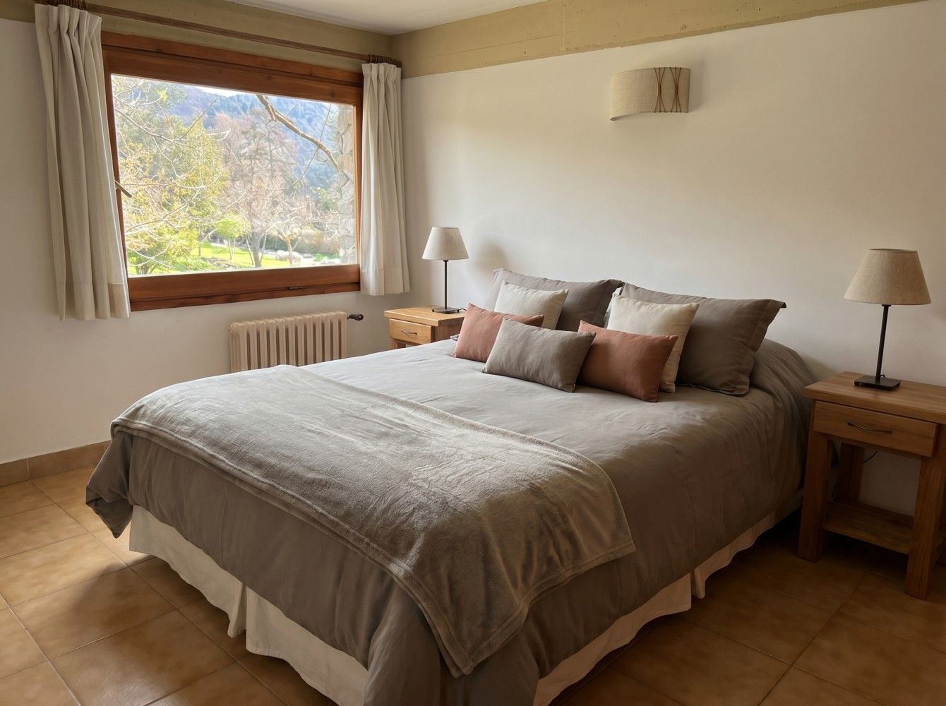 Habitación luminosa con una cama bien hecha, lámparas de noche y una ventana con cortinas y árboles otoñales en el exterior.