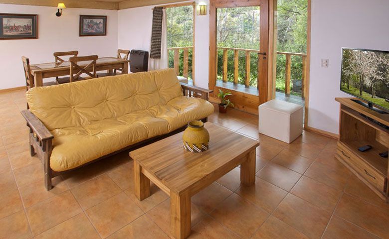 Luminoso salón con sofá de color beige, mesa de centro de madera, zona de comedor y puertas de cristal que dan a un jardín con vegetación.