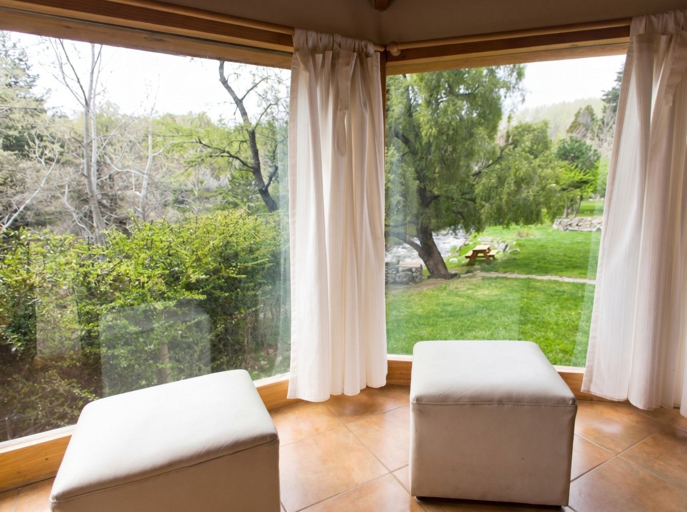 Habitación en esquina con ventanales del suelo al techo, cortinas blancas y dos otomanas blancas con vistas a un jardín verde.
