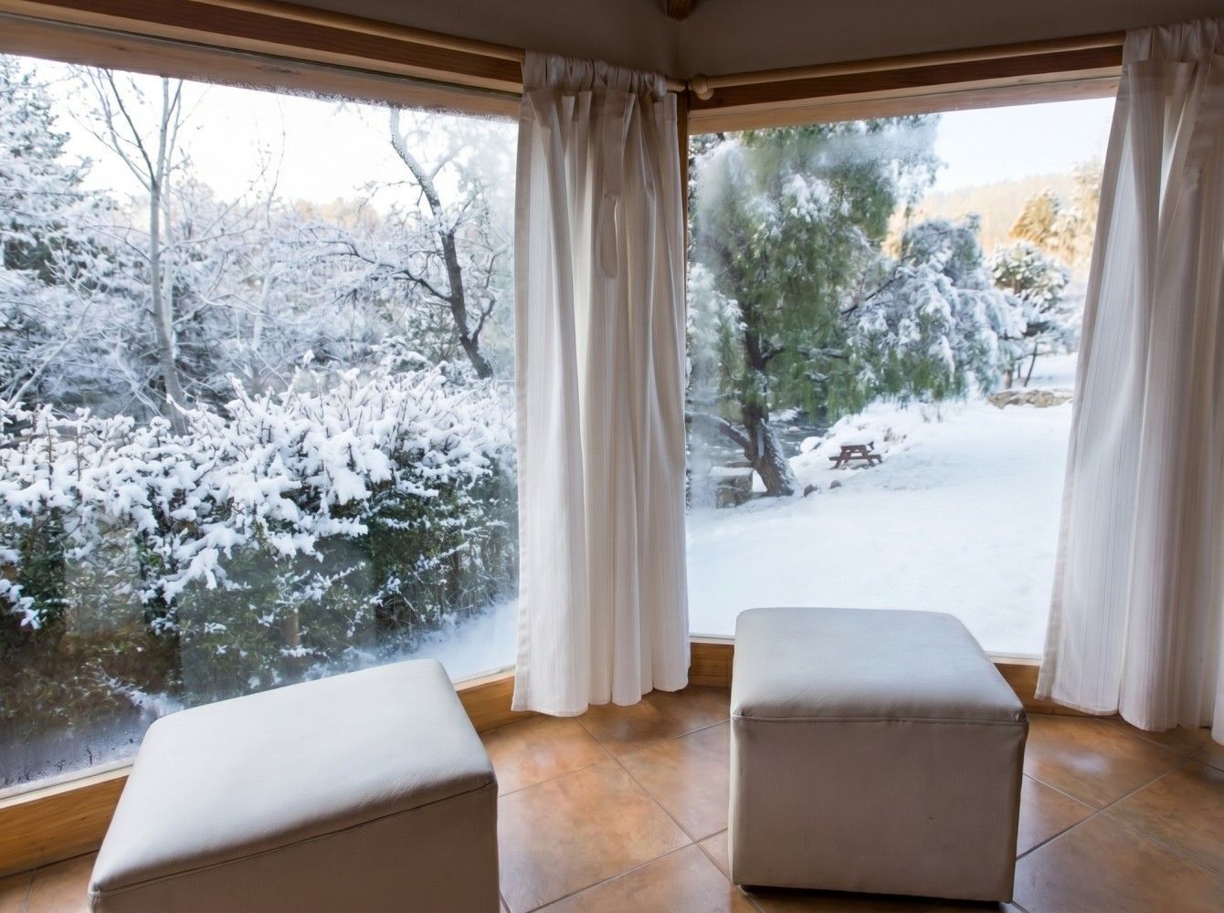 Vista invernal nevada a través de grandes ventanales desde una acogedora habitación con cortinas y cojines blancos.