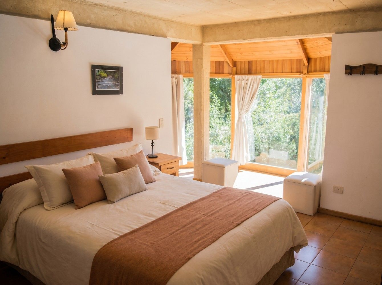 Habitación luminosa con cama, sábanas beige, cabecero de madera y grandes ventanales con vistas a los árboles.