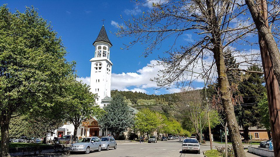 Plaza del pueblo bordeada de árboles, con una torre del reloj blanca bajo un cielo azul y coches aparcados a lo largo de la calle.