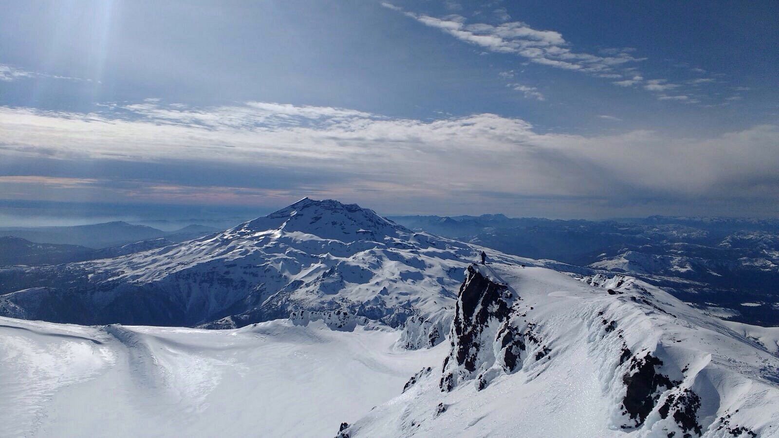 Cumbres nevadas bajo un cielo azul nublado, vistas desde una cresta elevada.