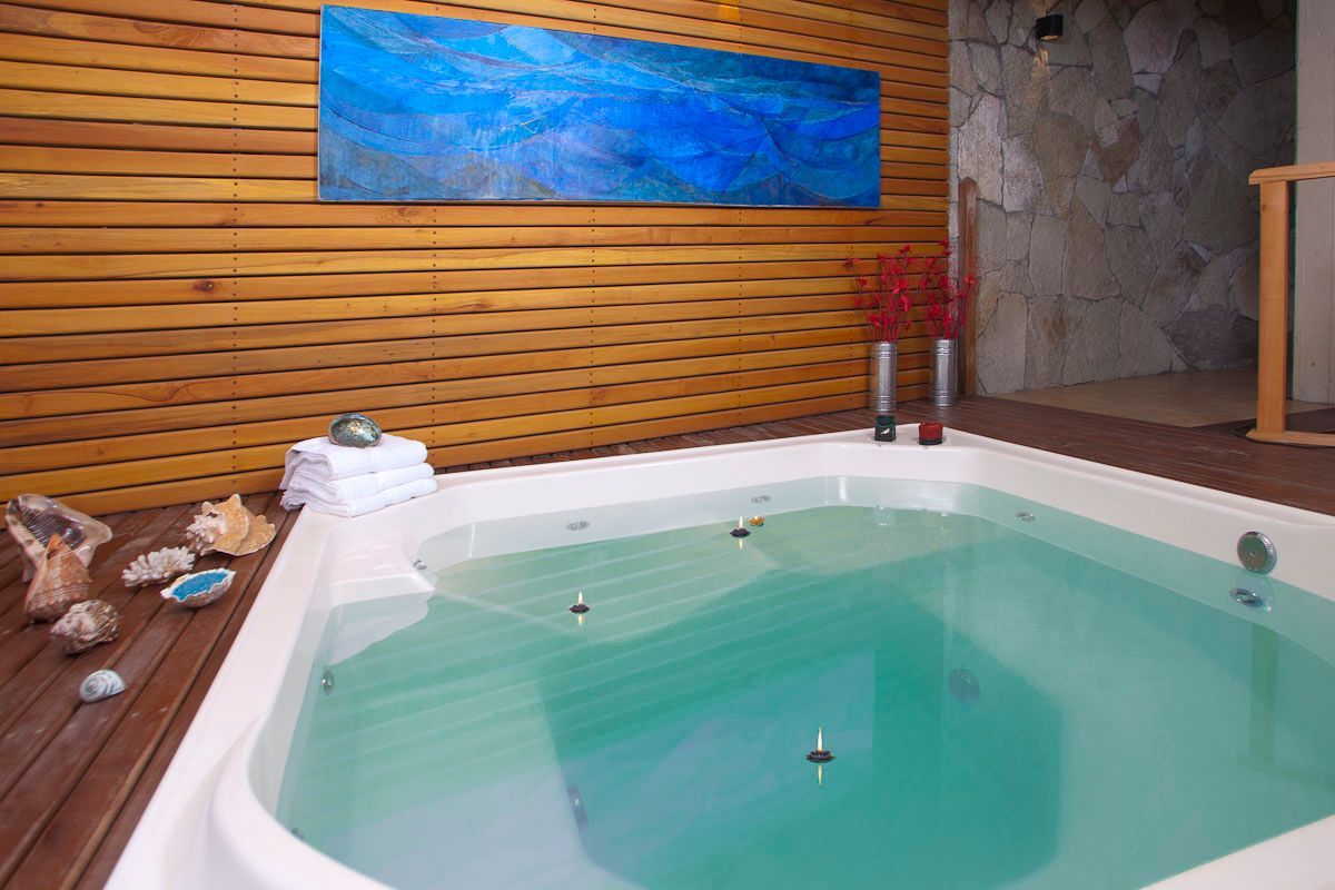 Bañera de hidromasaje interior con agua cristalina, pared revestida de madera, detalles en piedra y arte mural azul.