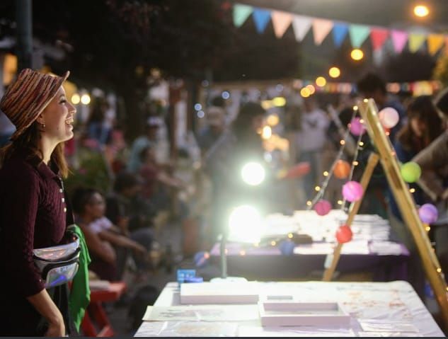 Una mujer con un abrigo granate está de pie en un puesto de un mercado nocturno, bajo coloridas guirnaldas y luces brillantes.