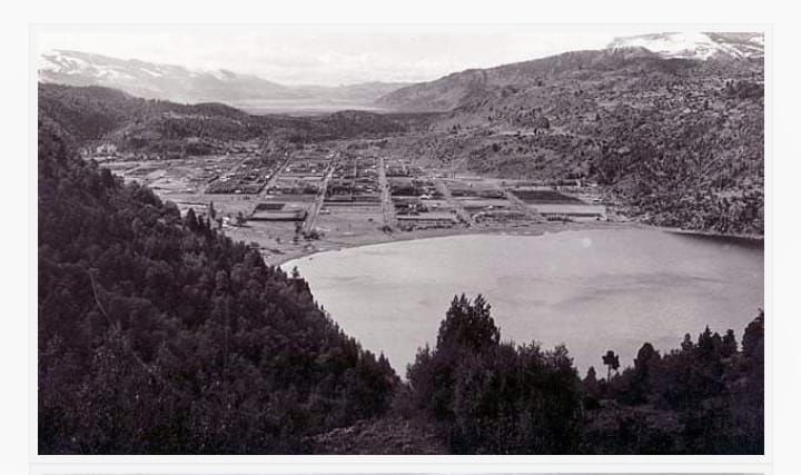 Vista en blanco y negro de un lago en un valle de montaña con campos, pueblos y colinas boscosas en primer plano.