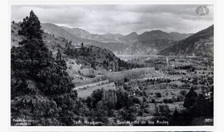 Valle de montaña en blanco y negro con una carretera sinuosa, edificios dispersos y un río en la distancia.