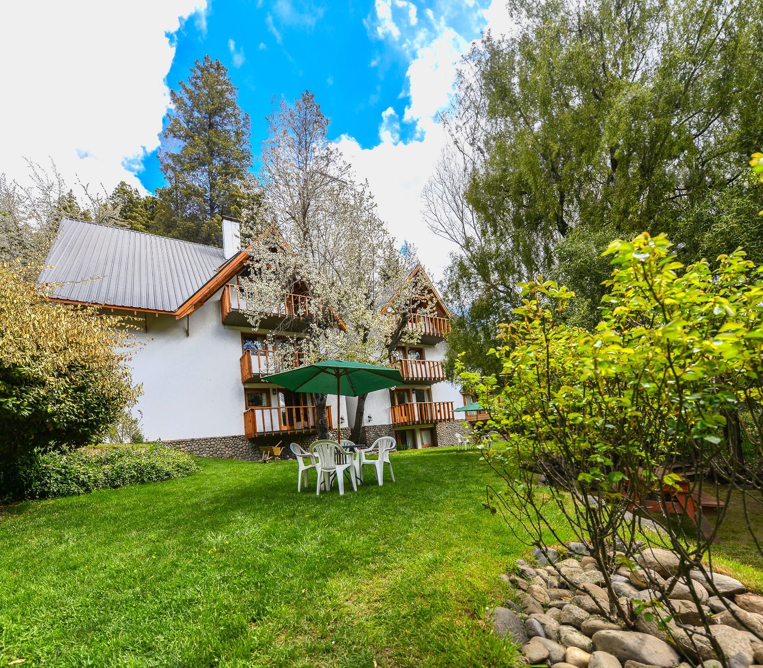 Casita blanca con sombrilla verde en el patio, sillas blancas y un jardín exuberante bajo un cielo azul.