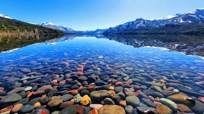 Un lago de montaña cristalino con rocas coloridas bajo aguas azules tranquilas y picos nevados reflejados en la superficie.