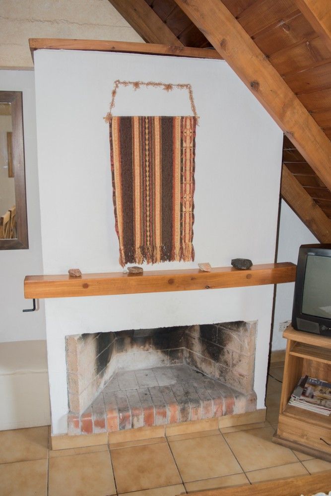 Acogedora chimenea con repisa de madera, tapiz de pared a rayas y televisor en una habitación con techo inclinado.