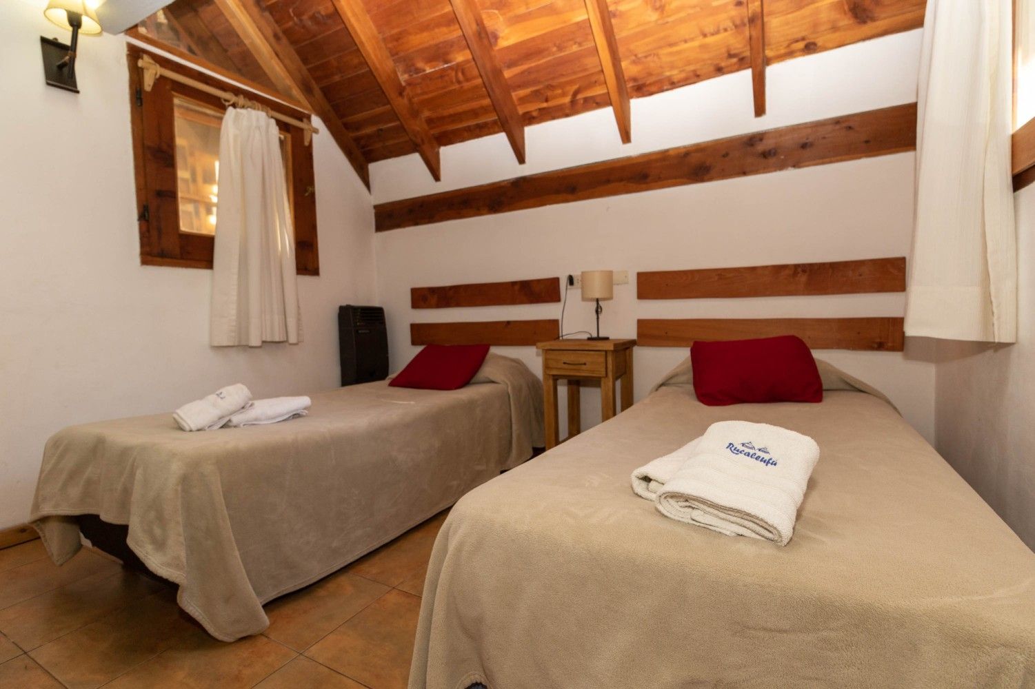 Habitación pequeña de estilo rústico con dos camas individuales, paredes blancas, techo de madera e iluminación cálida.