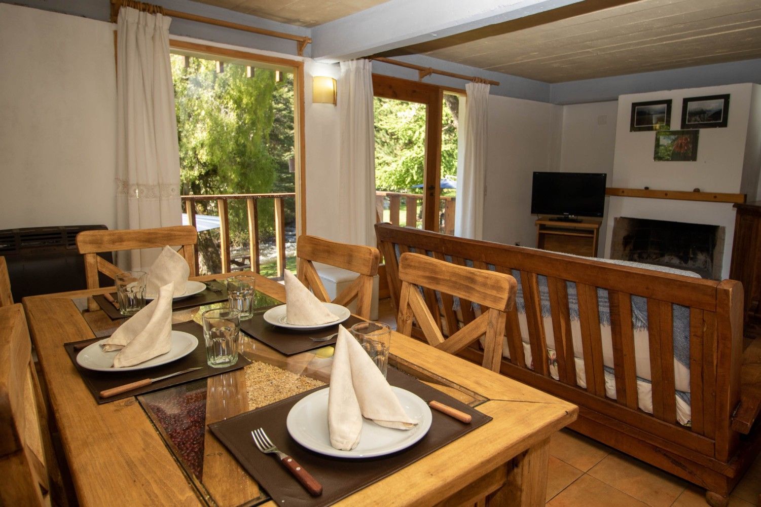 Comedor de madera con mesa puesta, puertas abiertas que dan al balcón y una chimenea al fondo.