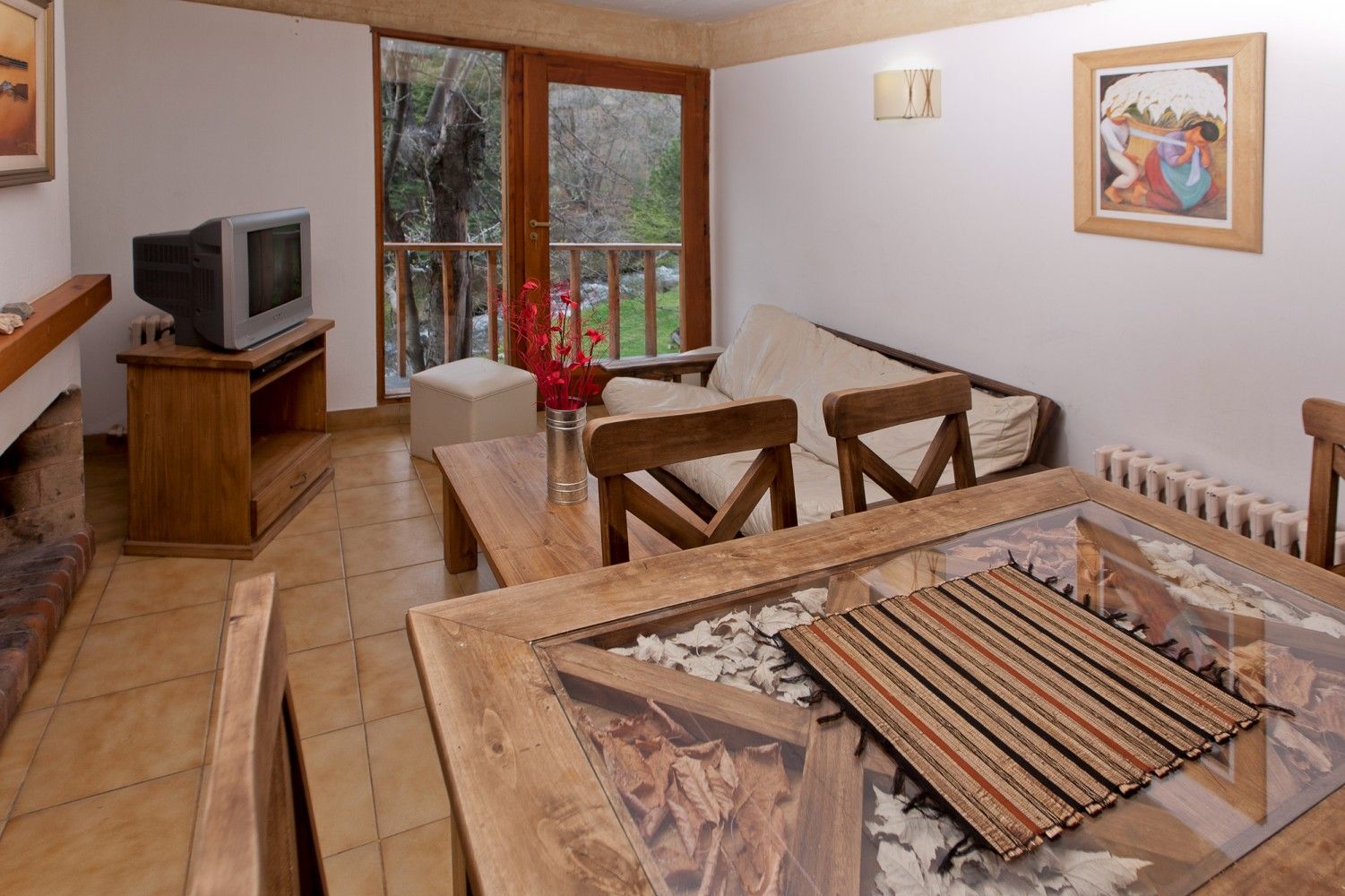 Acogedora sala de estar con televisión, asientos de mimbre, mesa de madera y puertas que dan a un balcón con vegetación.