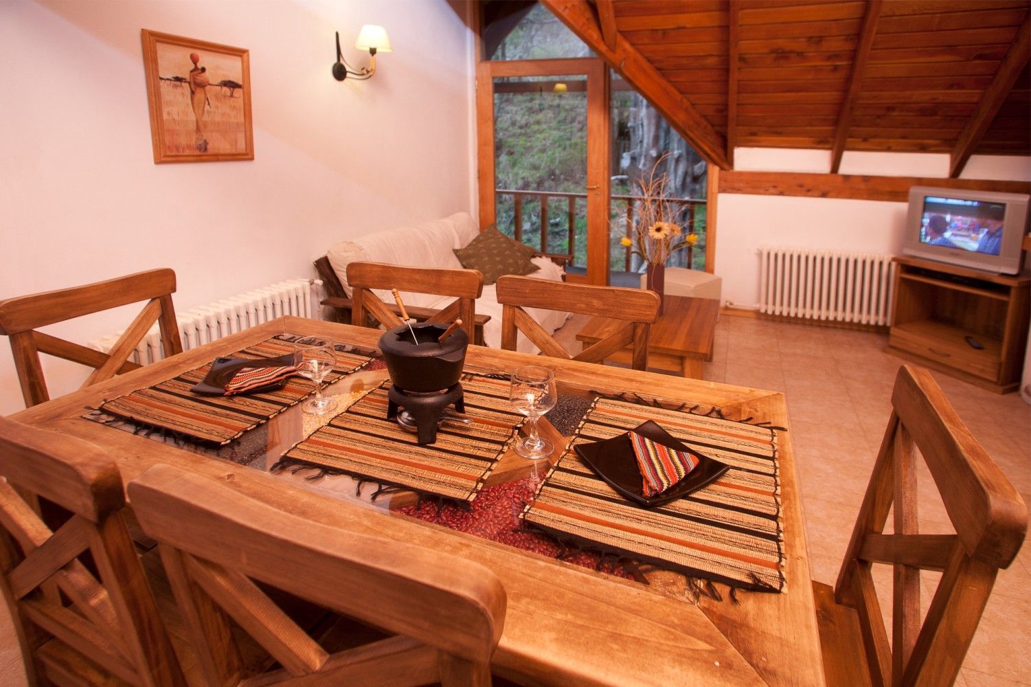 Acogedor comedor de madera con mesa puesta, grandes ventanales y televisor sobre un mueble.