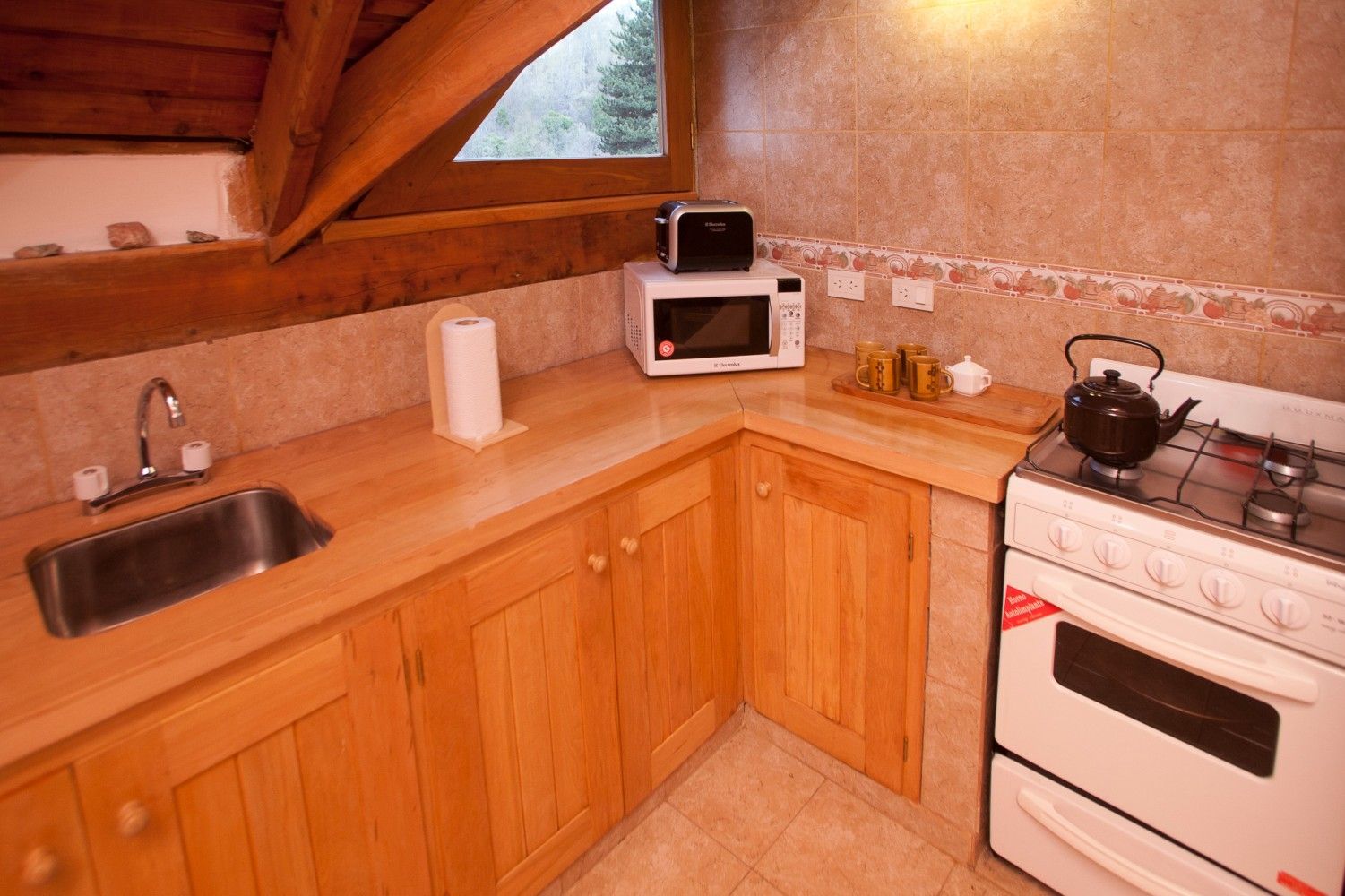 Pequeña cocina rústica con armarios de madera, fregadero, microondas, fogones y una ventana bajo un techo inclinado.