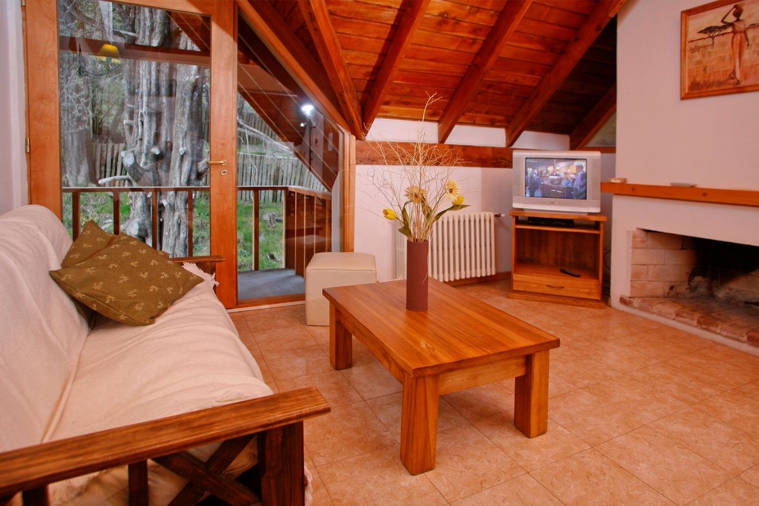 Acogedora sala de estar con techo de madera, sofá blanco, mesa de centro, chimenea, televisor y puertas que dan a un balcón con vistas a los árboles.