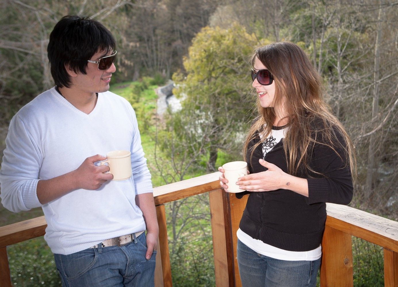 Dos personas con tazas en la mano y sonriendo en una terraza de madera con una ladera boscosa al fondo.