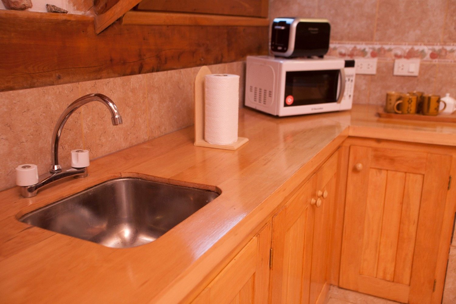 Encimera de cocina compacta con fregadero de acero inoxidable, armarios de madera, rollo de papel de cocina y microondas blanco.
