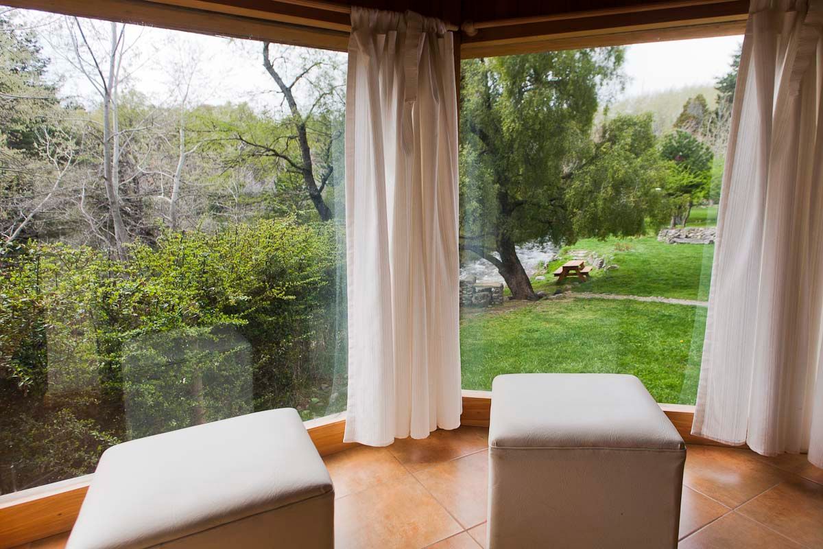 Patio cubierto con cortinas color crema, otomanas y vistas a un jardín verde y árboles junto a un estanque.