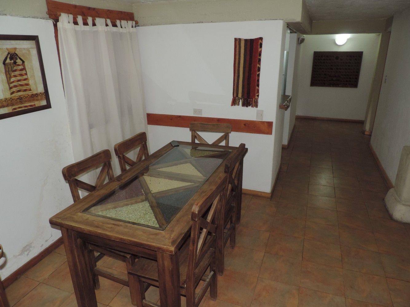 Pasillo interior con poca luz, con una mesa y sillas de madera junto a paredes blancas y molduras rojas.