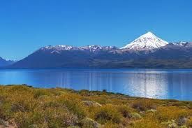 Montaña cubierta de nieve sobre un tranquilo lago azul, con una orilla cubierta de hierba en primer plano.