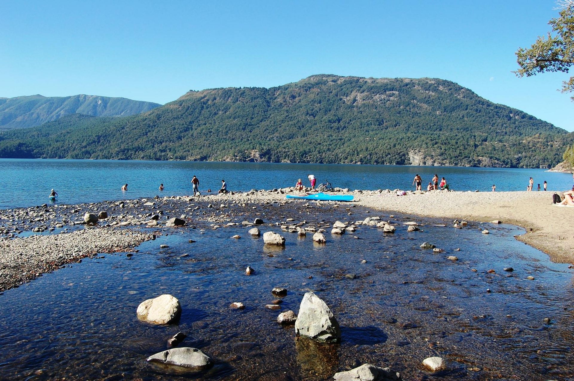 Playa de guijarros en un tranquilo lago azul con gente nadando y montañas verdes al fondo.