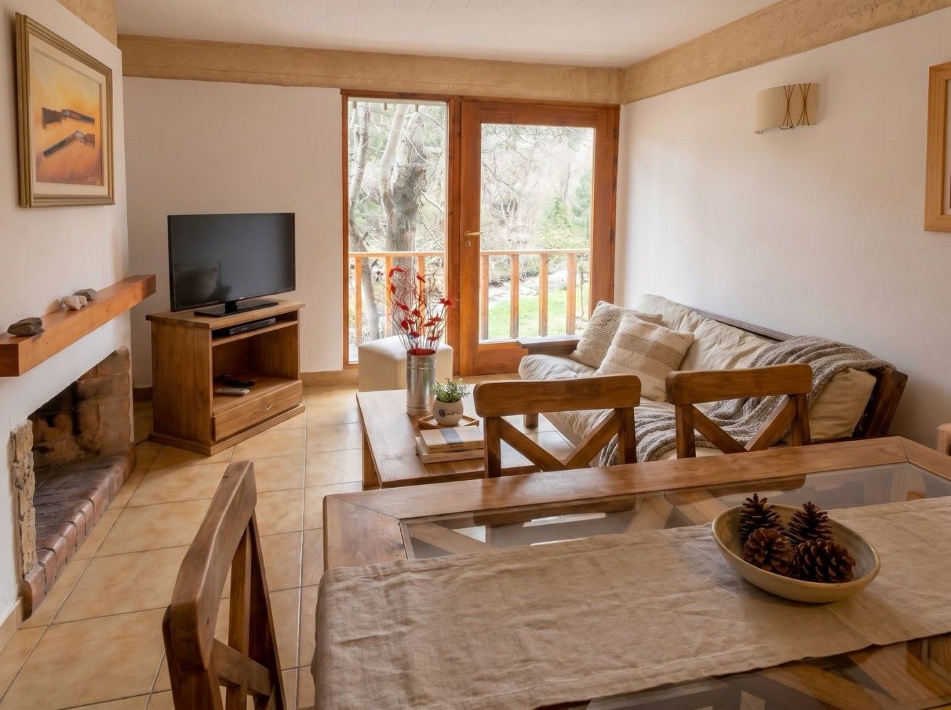 Acogedora sala de estar con sofá, televisor, mesa de madera, chimenea y puertas que dan a un balcón con vistas a los árboles.
