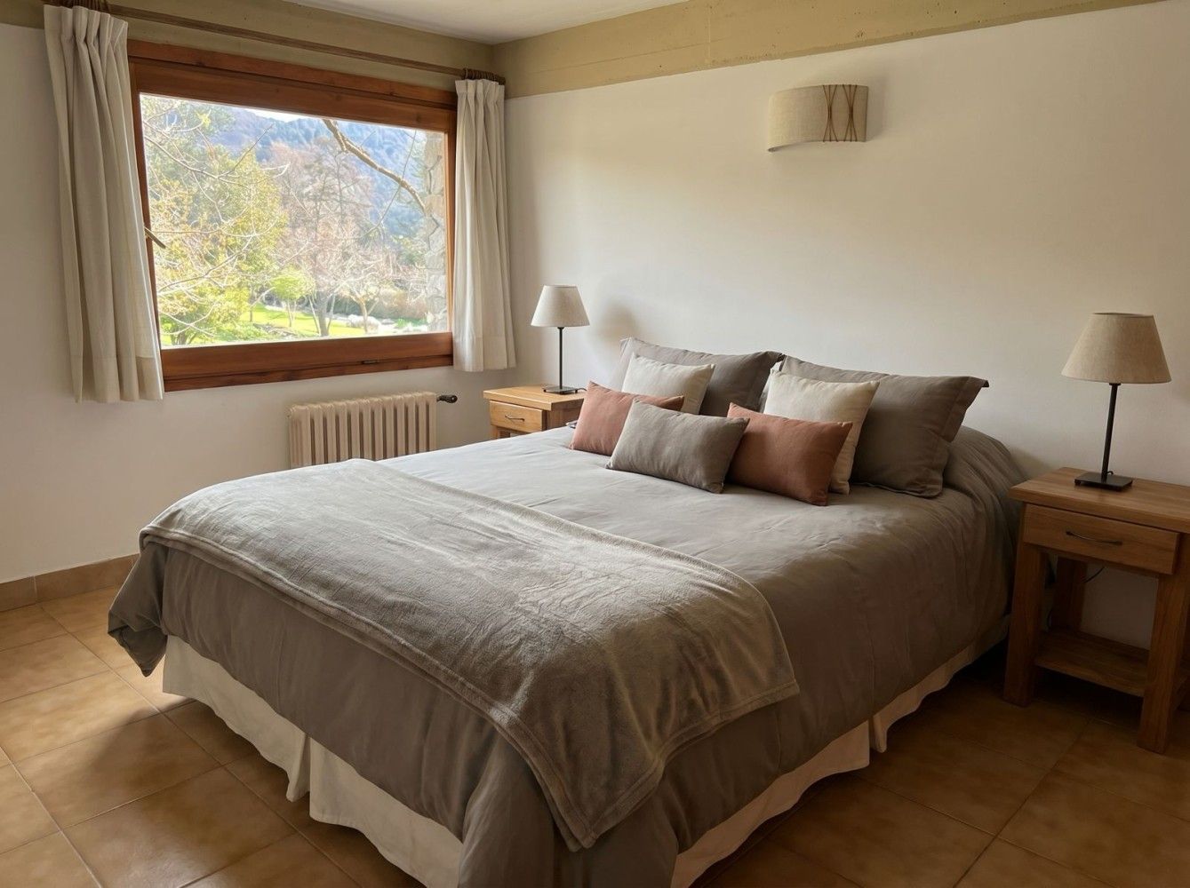 Habitación luminosa con una cama bien hecha, paredes de color beige, dos lámparas de noche y una ventana con cortinas.