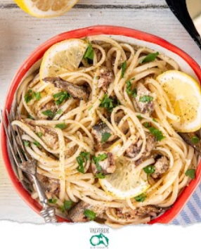 Una ciotola rossa di spaghetti con funghi, fette di limone e prezzemolo su una tovaglia chiara.