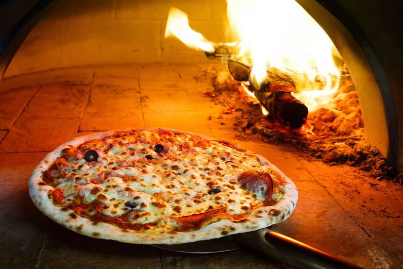 Pizza in forno a legna, con le fiamme a destra e una pala per pizza sotto.