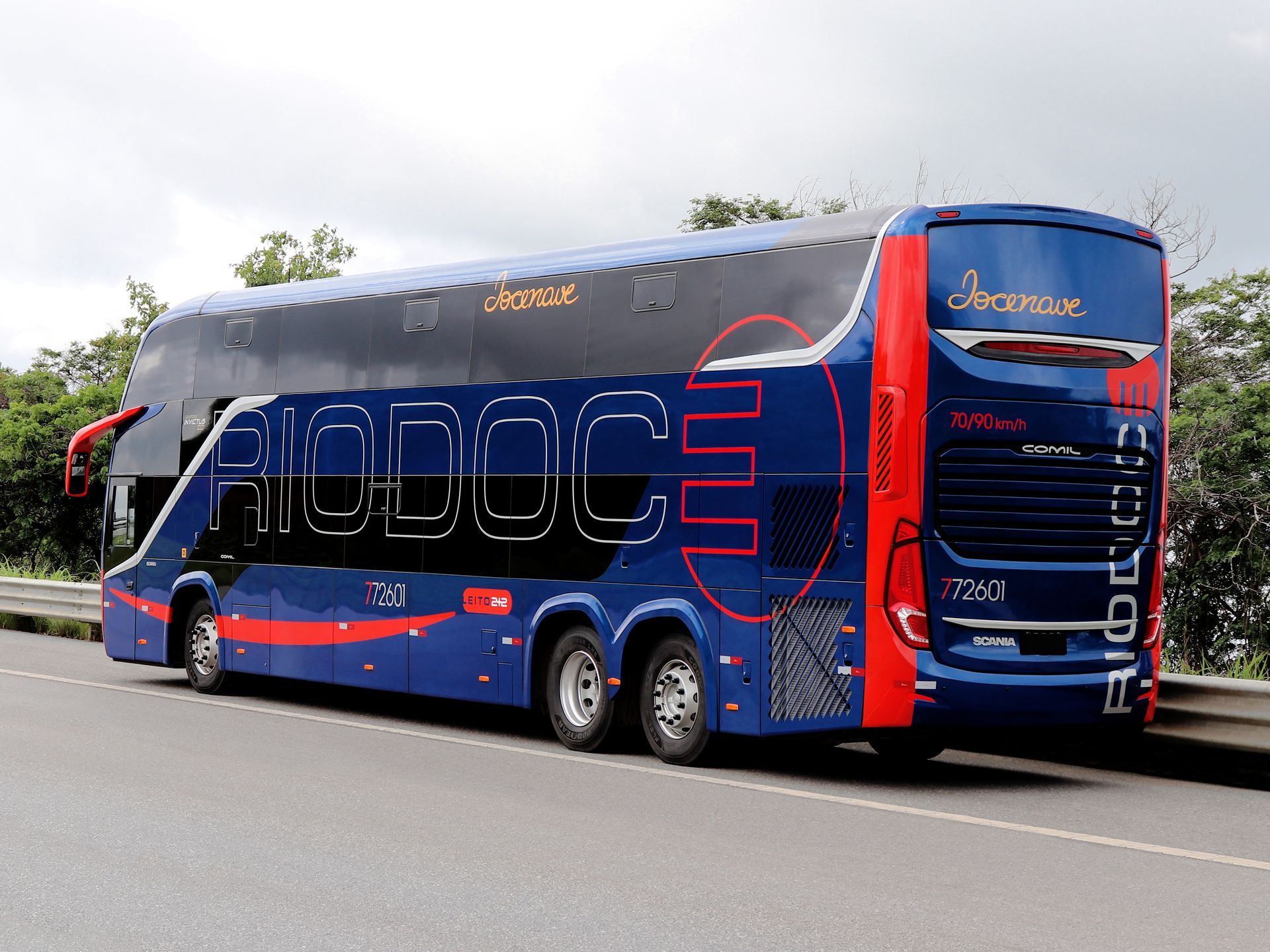 Um ônibus de dois andares Rio Doce, azul e vermelho, estacionado na beira da estrada com árvores ao fundo.
