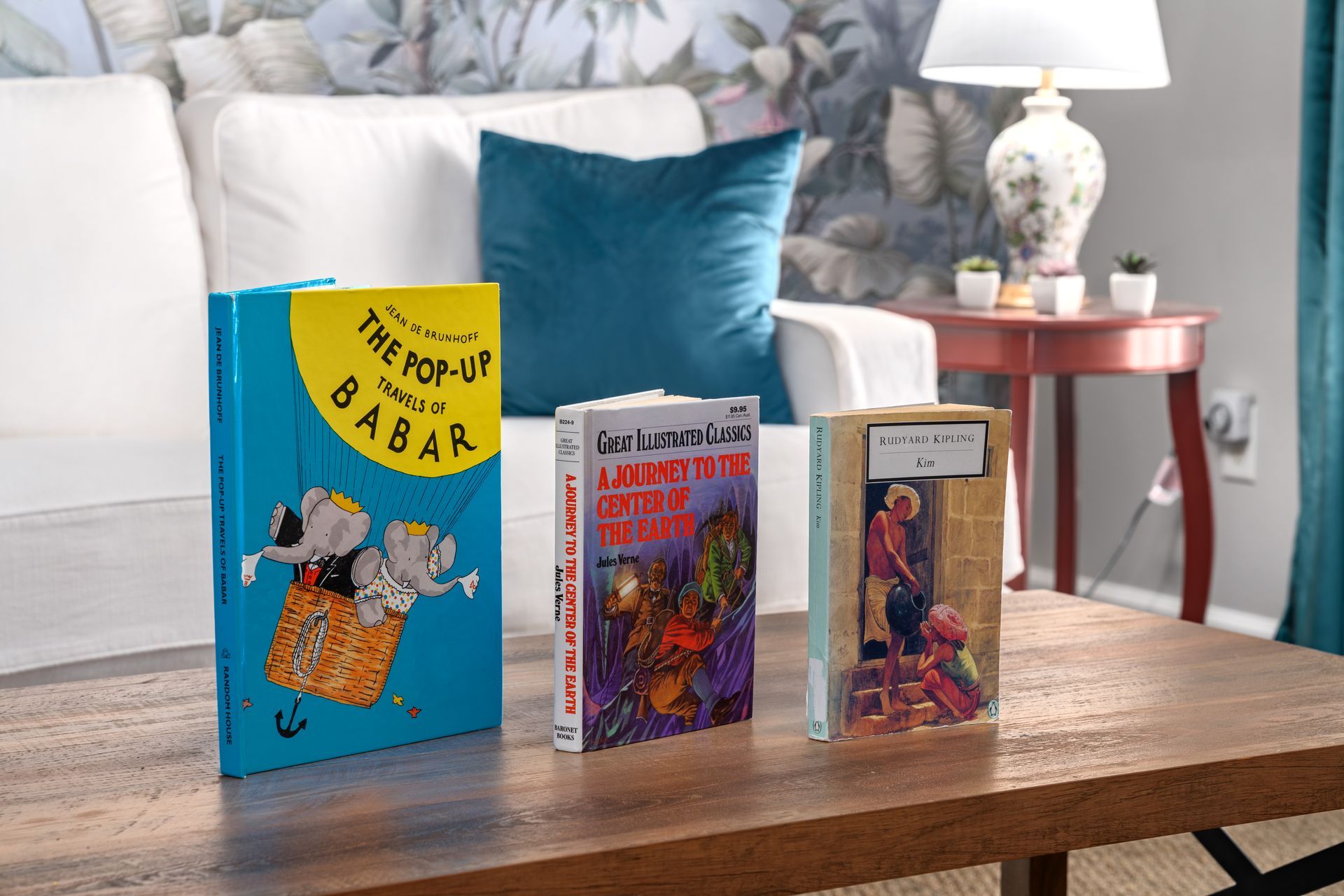 Three books displayed on a wooden table in a living room, with a white sofa and lamp in the background