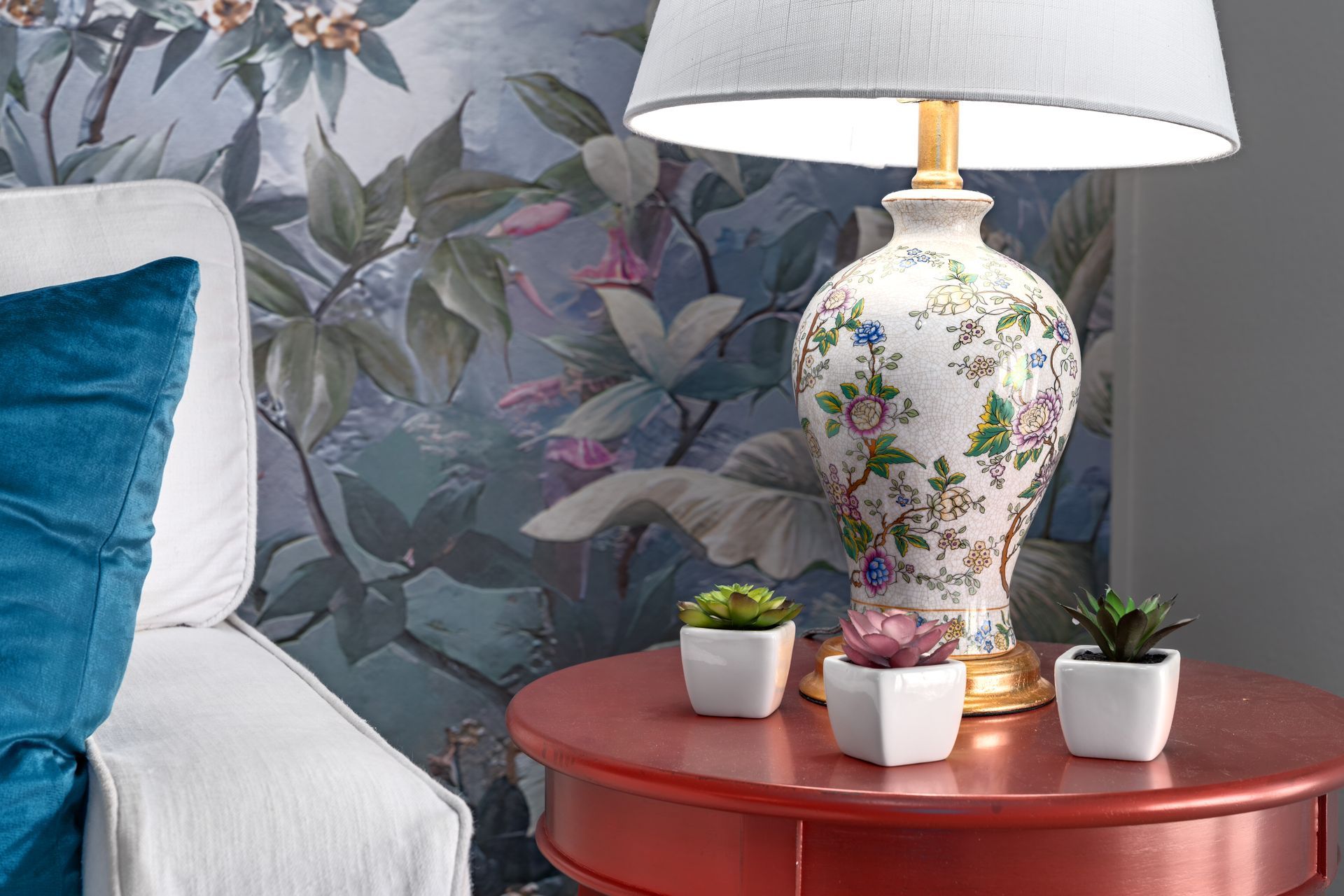 Floral lamp on a red side table with small potted succulents beside a white chair and blue floral wallpaper.