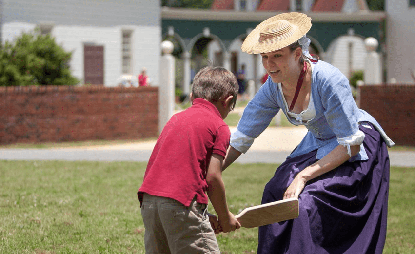 Woman in colonial dress teaches boy to play paddle ball outdoors.