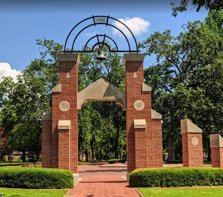 Red brick archway with a bell, set in a tree-lined walkway, under a blue sky.