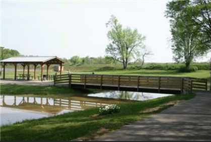 Wooden bridge over a pond in a park, with a pavilion in the background.