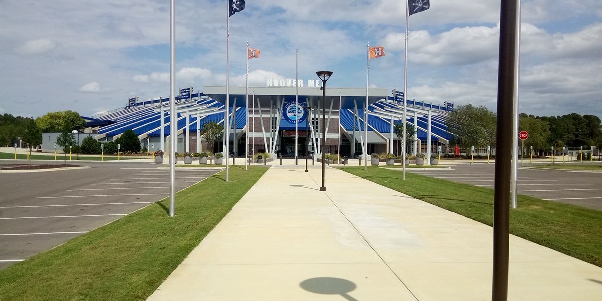 Exterior view of a large blue and white stadium, a pathway in the foreground, and flags waving on a sunny day.