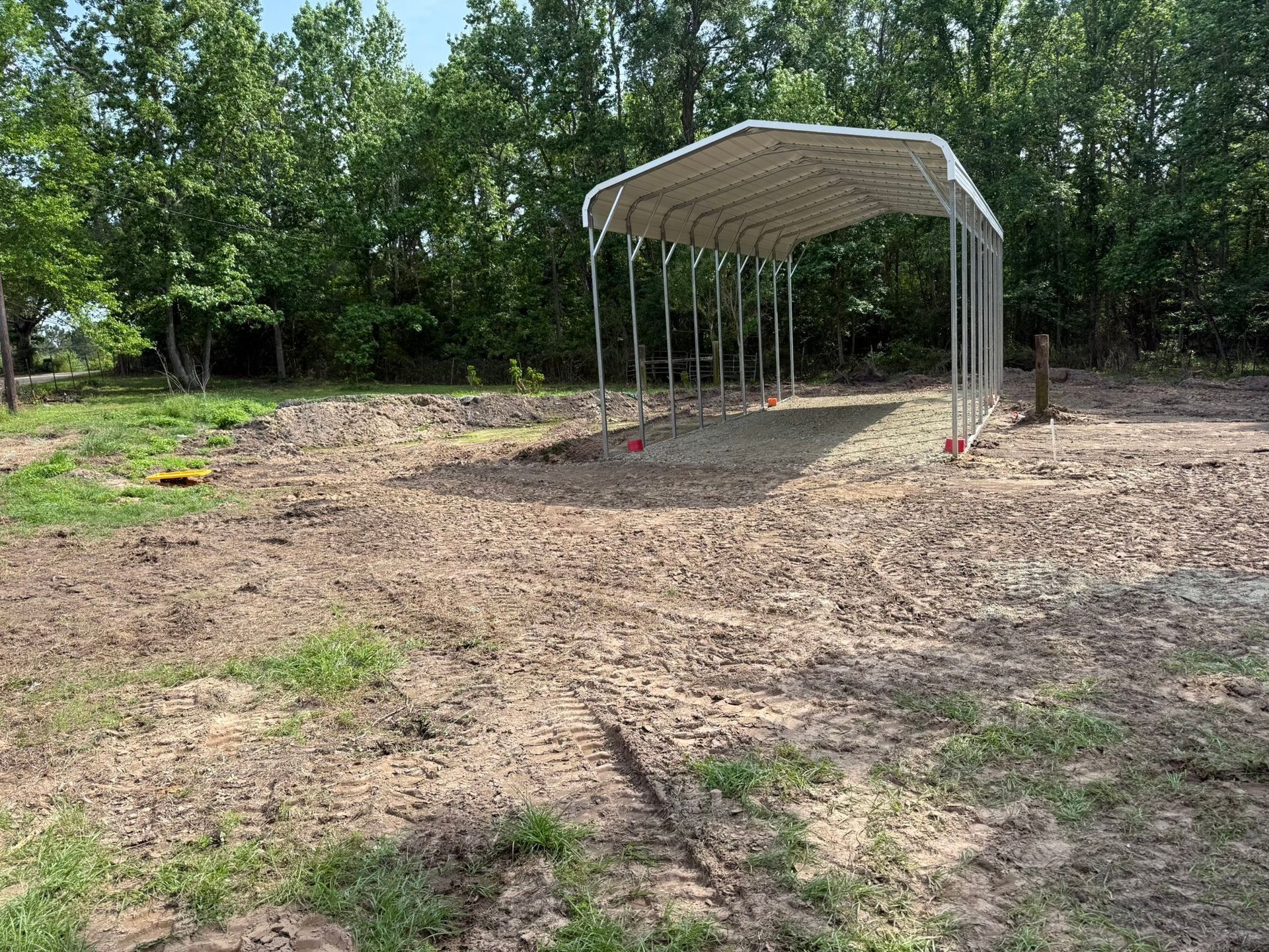 Empty dirt lot with a metal-roofed carport frame near trees and grass
