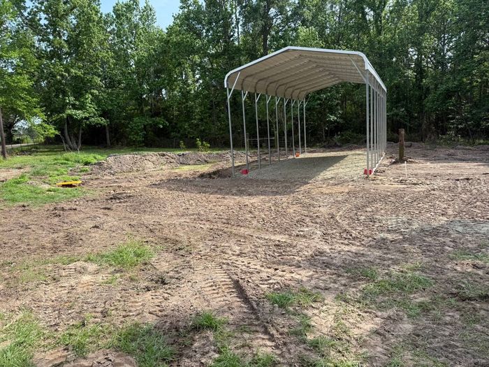 Empty dirt lot with a metal-roofed carport frame near trees and grass