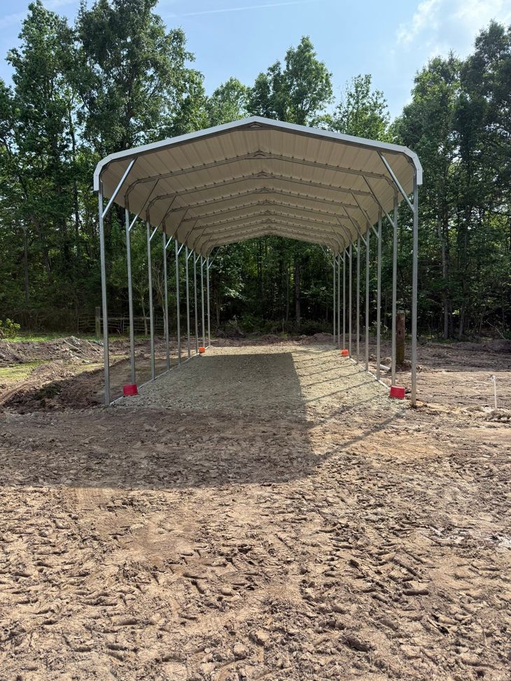 Open-sided metal carport with a white roof on a dirt lot, surrounded by trees